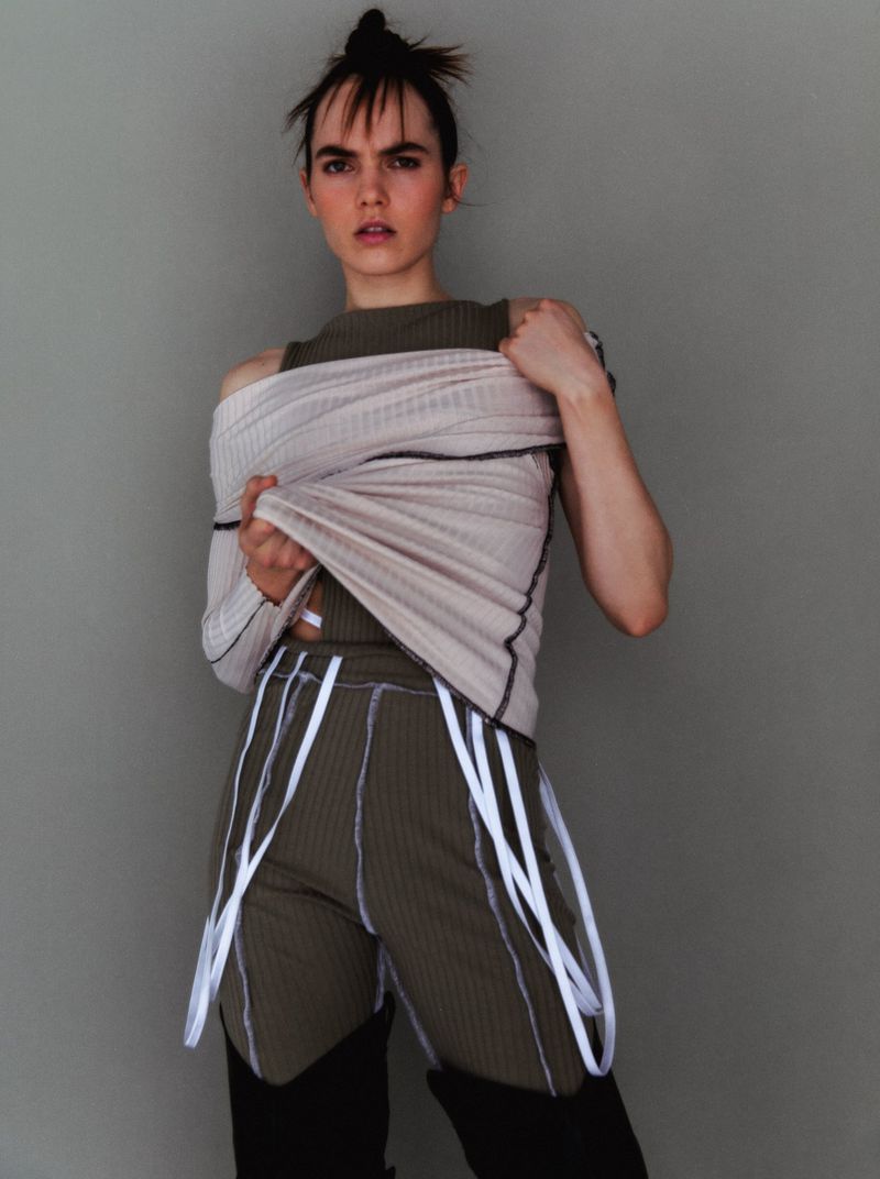 Fashion-forward portrait of a young adult woman in a beige and olive green ribbed top and shorts with white accents, against a grey wall.