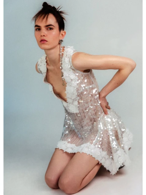 Young adult female model kneeling in a white sequin dress with floral detailing, set against a light blue-grey backdrop in a fashion editorial.