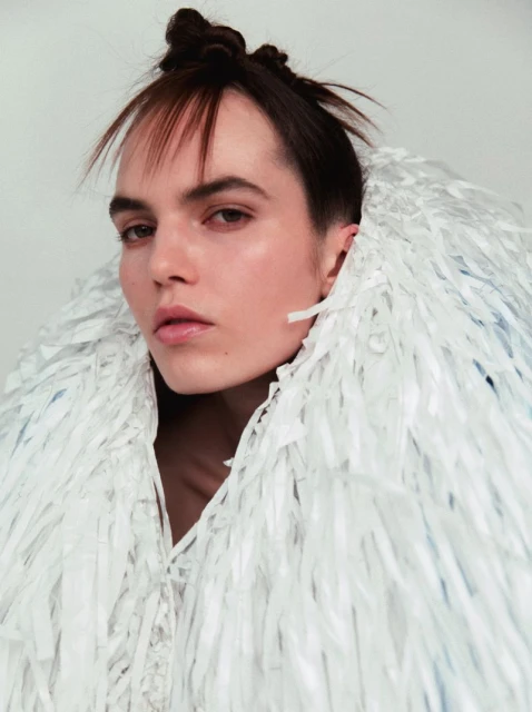 Close-up portrait of a woman with dark hair, styled in a unique bun, wearing a decorative white fringe collar against a plain white background.