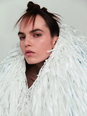 Close-up portrait of a woman with dark hair, styled in a unique bun, wearing a decorative white fringe collar against a plain white background.