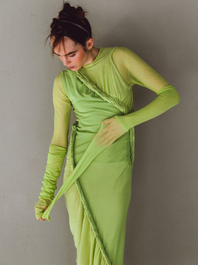 A woman poses in a light green dress with braided accents, creating a soft, ethereal look against a muted grey backdrop in a studio setting.