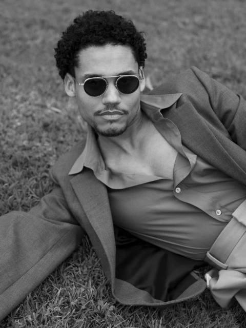 A man with curly hair and aviator sunglasses poses in an unbuttoned shirt and jacket on a grassy field in a black and white portrait.