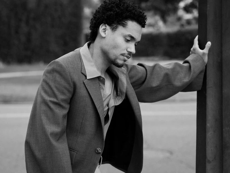 Black and white portrait of a young adult man in a blazer jacket, leaning against a pole outdoors, with blurred background. Fashion photography.