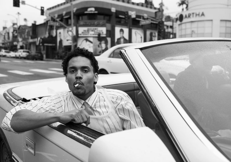 A man with a cigarette poses in a convertible car in a black and white outdoor shot with buildings and traffic in the background.