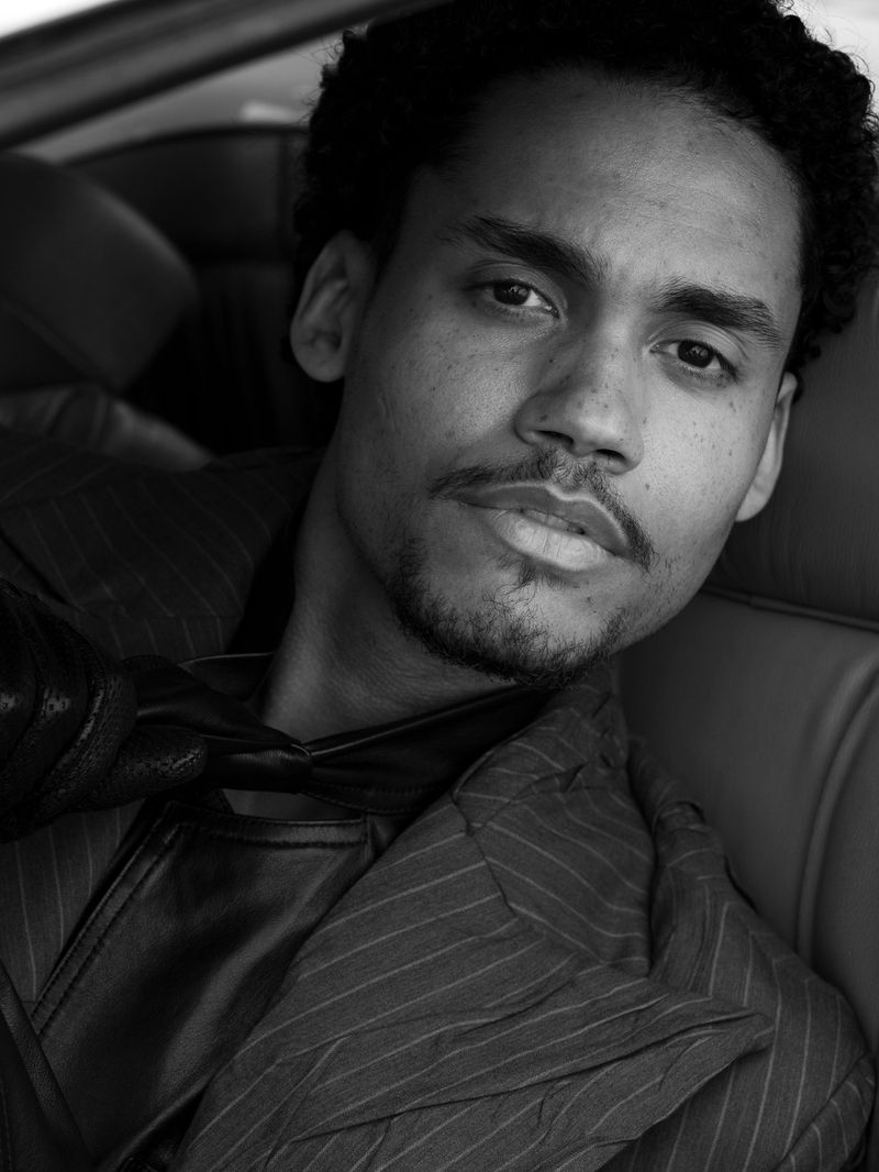 Black and white portrait of a man with dark curly hair, wearing a pinstripe jacket and leather vest, sitting in a car. 