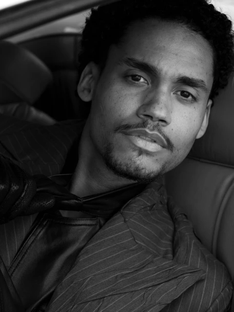 Black and white portrait of a man with dark curly hair, wearing a pinstripe jacket and leather vest, sitting in a car. 