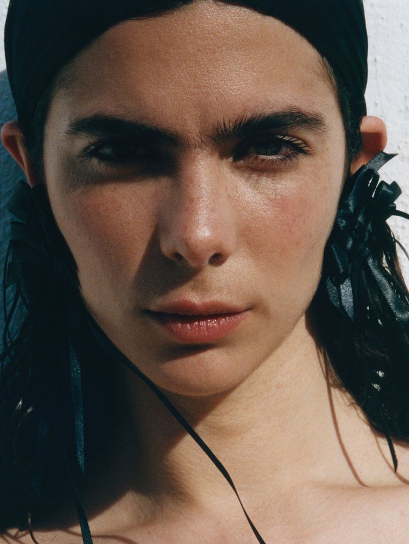 Close-up portrait of a woman with dark hair and eyebrows, fair skin, wearing a black headscarf and dark ribbon earrings against a white wall.