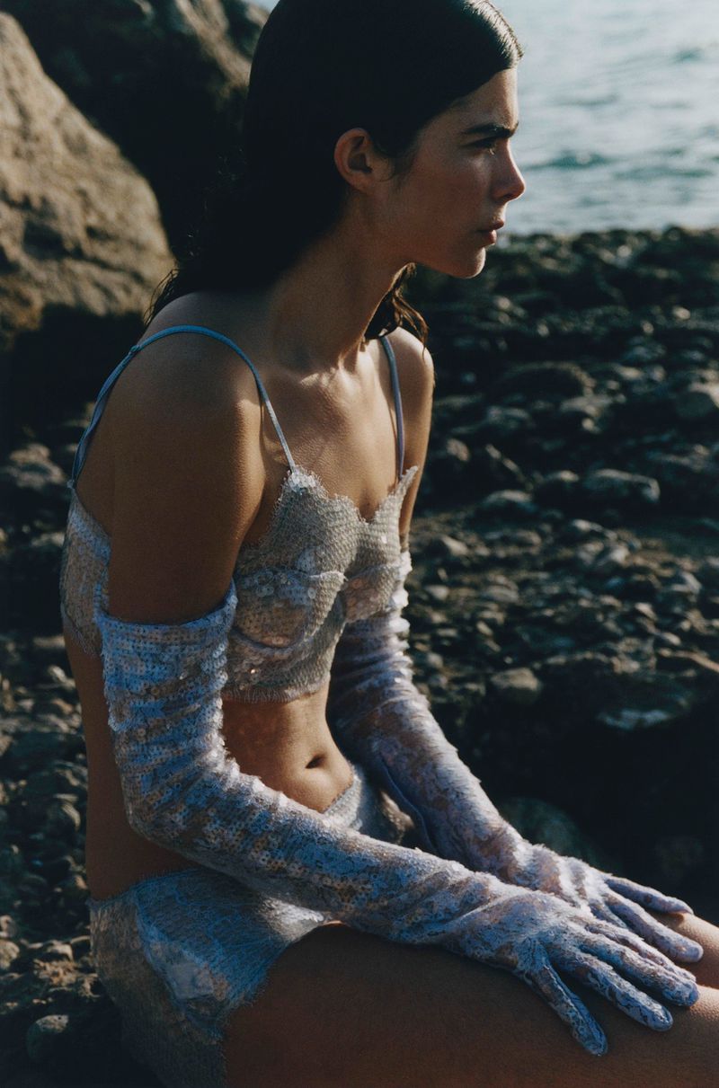 Woman in a shimmering silver and light blue two-piece outfit with long gloves sitting on rocks by the water looking off to the side.