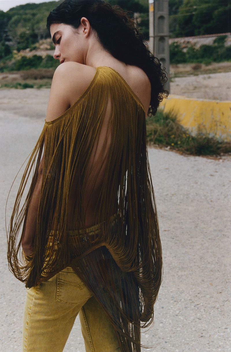 Woman in yellow jeans and fringed top stands on a road with a natural backdrop featuring vegetation and power poles.