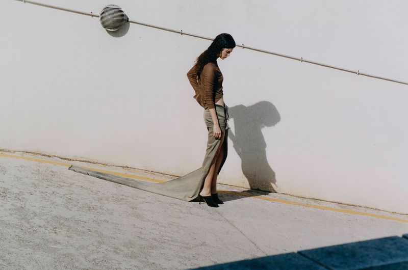 Brunette woman posing in olive green slit skirt and brown top with white wall and concrete floor in an outdoor setting.