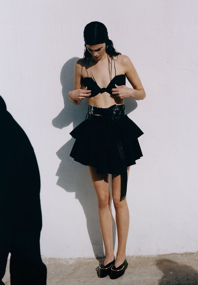 Young woman poses against a white wall in black two-piece fashion. She wears a headscarf, a two-tier skirt, and platform shoes.
