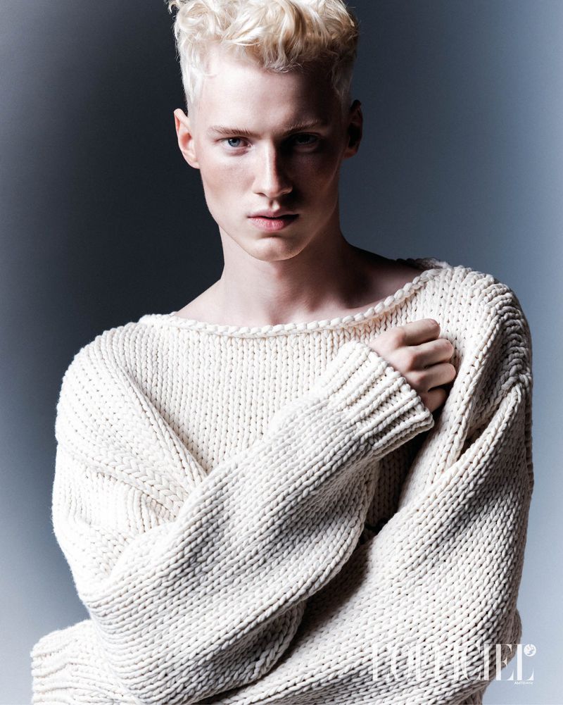 Portrait of a fair-skinned, blonde-haired person wearing a knitted sweater, set against a soft grey backdrop in a close-up studio shot.
