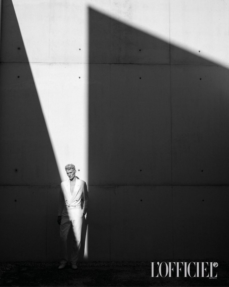 Black and white portrait of a male fashion model in white suit, dramatically lit against a concrete wall, showcasing L'Officiel magazine logo.