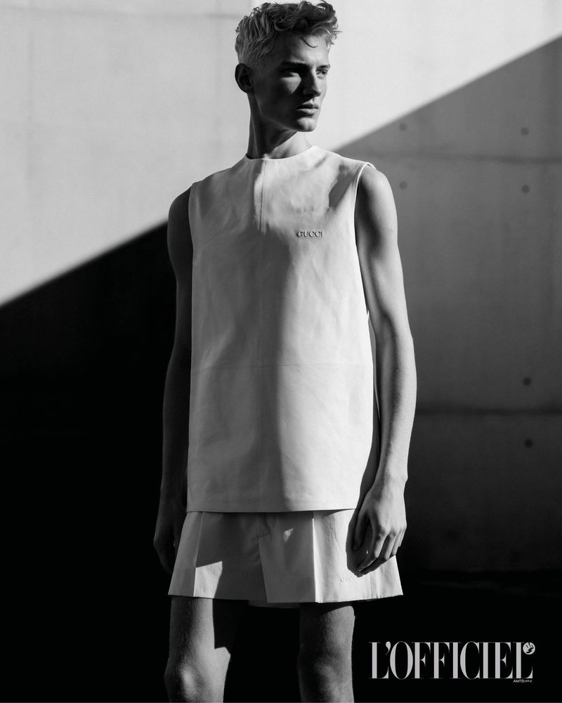 Black and white portrait of a male model wearing a sleeveless top and shorts against a concrete wall with dramatic harsh shadows.