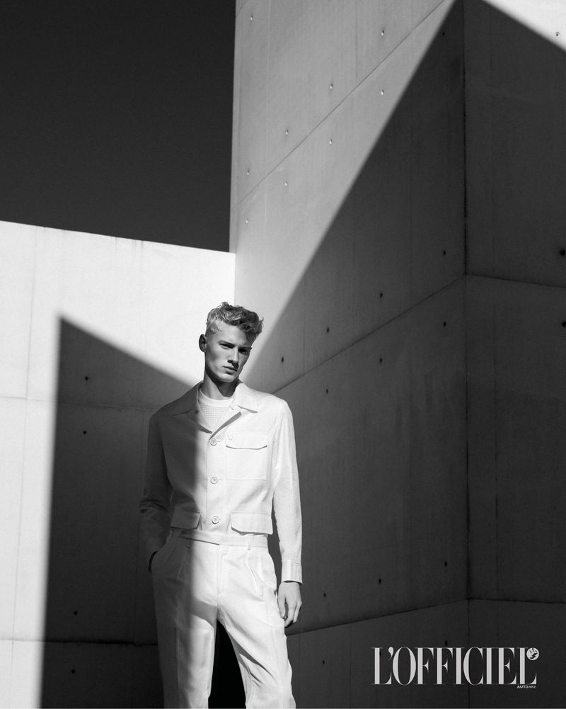 Monochrome fashion shot of a blond male model in a light-colored suit, posing against a textured concrete wall with geometric shadows.