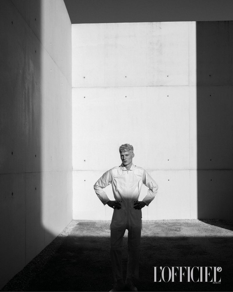 Black and white portrait of a blond man in a light-colored jumpsuit standing in a concrete architectural space with hands on hips.