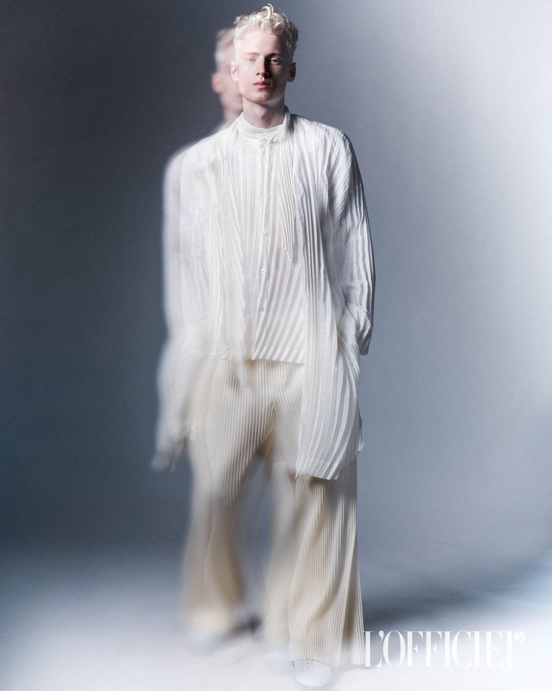 Man with platinum blonde hair posing in a studio wearing a textured white long-sleeved shirt and flowing pants in soft, diffused lighting.