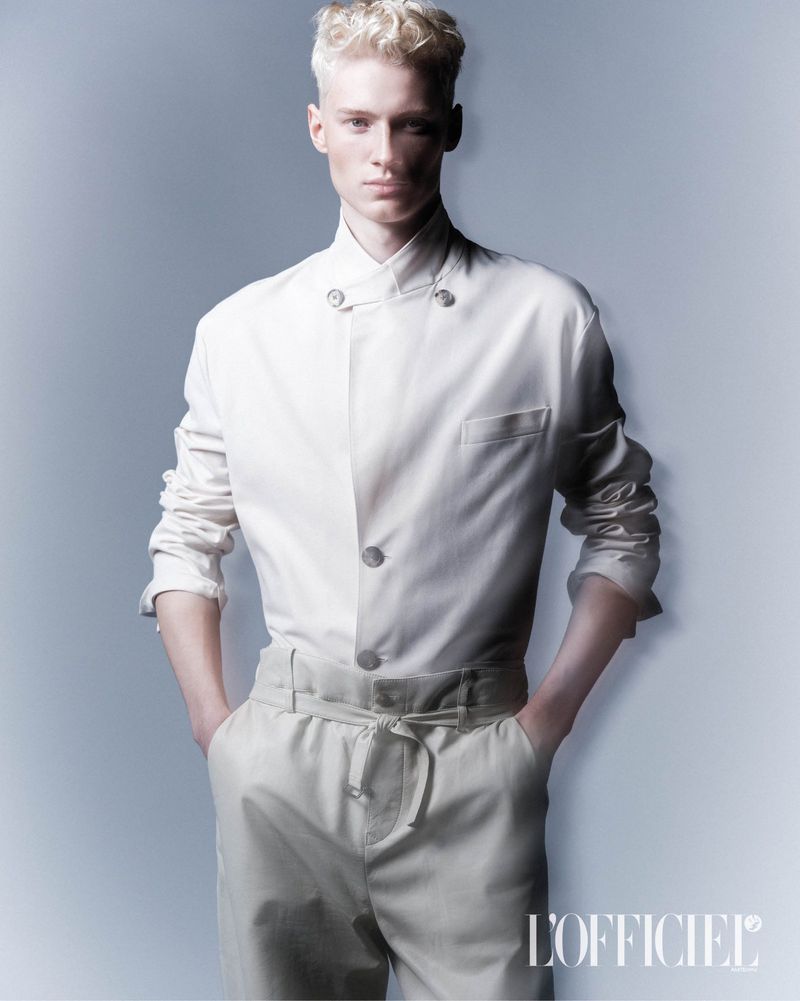 Fashion portrait of a blonde male model in a white button-down shirt and light pants, posing in front of a soft blue-grey backdrop.