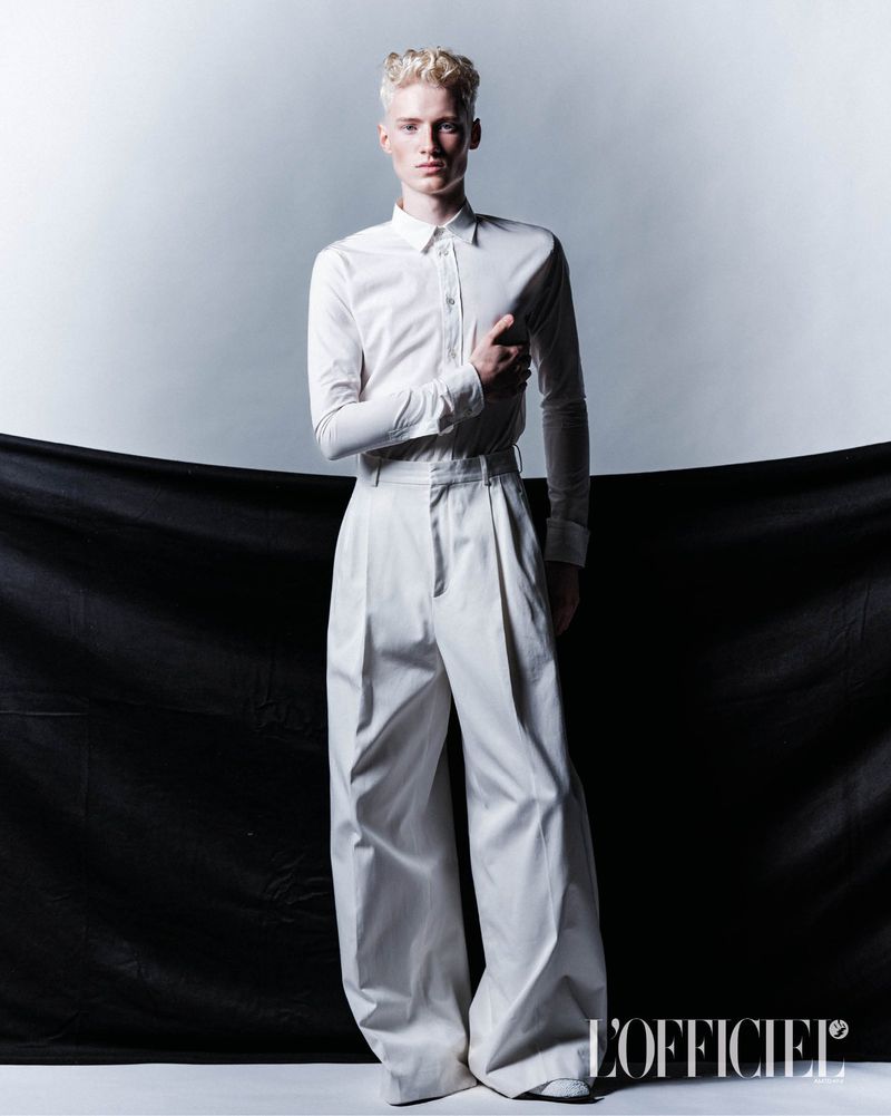 Full-length studio shot of a male fashion model with platinum blond hair, wearing a white collared shirt and high-waisted, wide-leg trousers.