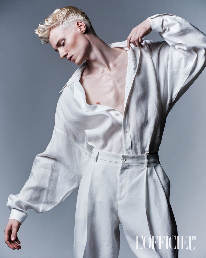 Blond male fashion model poses in an unbuttoned white linen shirt and matching trousers against a neutral grey background.