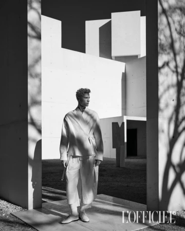 Monochrome portrait of a blonde man in a kimono-style top and pants stands in front of a modern building with tree shadow.
