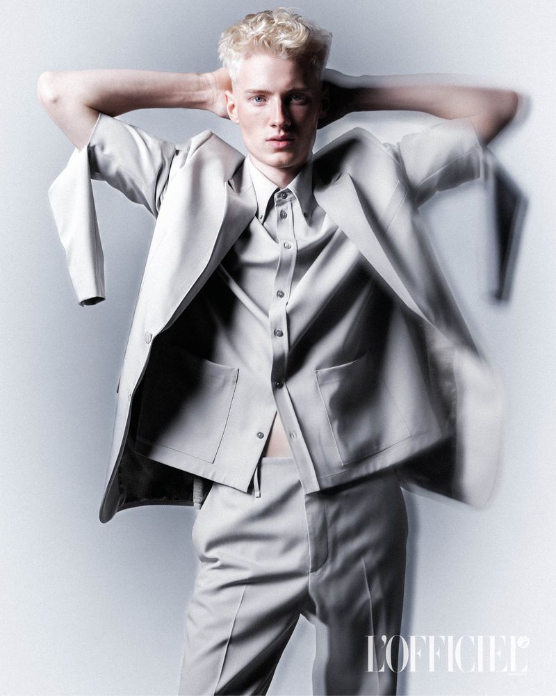 Fashion shot of a light-skinned blonde male model wearing a light gray suit. He is posed in a photo studio against a light background.