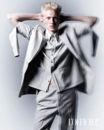 Blonde male model wearing a light grey suit with hands behind head against a white seamless background