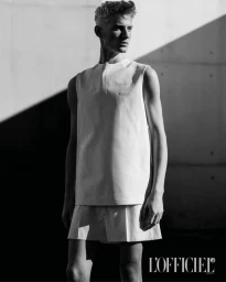 Black and white portrait of a male model wearing a sleeveless top and shorts against a concrete wall with dramatic harsh shadows.