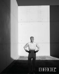 Model with light hair standing in a white jumpsuit inside a tall concrete courtyard with strong shadows for L'Officiel Hommes Italia.