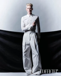 Blonde male model wearing a white buttoned shirt and wide trousers against a studio background with a black fabric drape