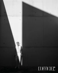 Model wearing a white double-breasted suit standing against a dark concrete wall with dramatic sharp diagonal shadows.