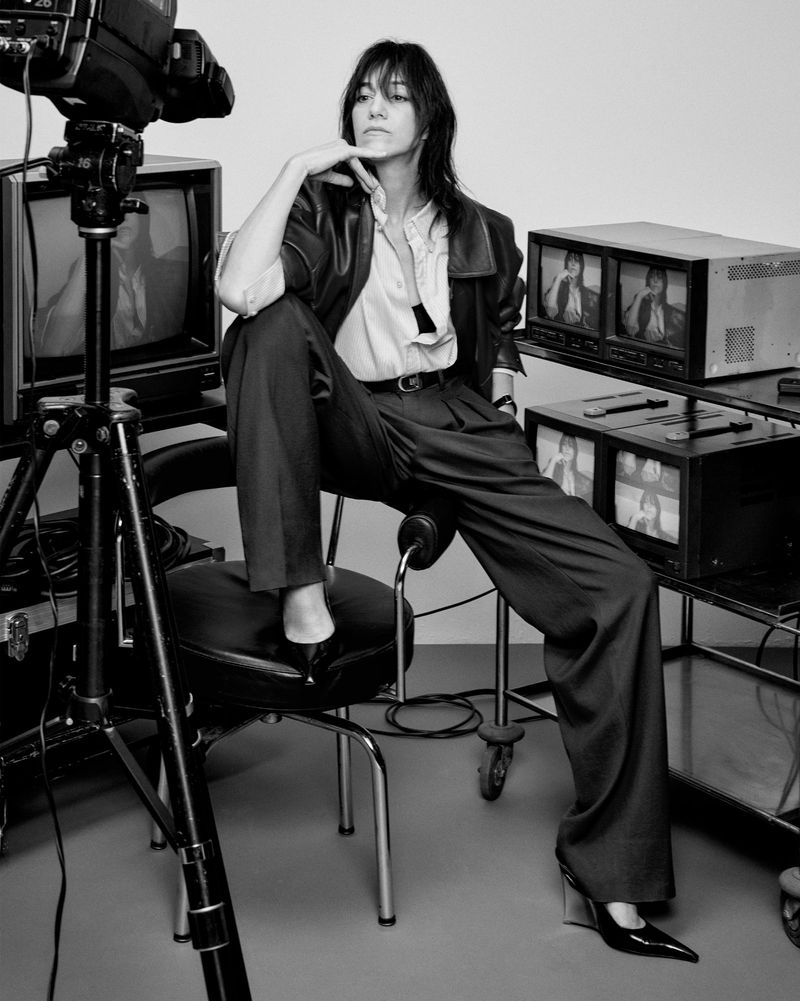 Black and white studio shot of a woman in a pantsuit sitting on a chair surrounded by vintage television equipment and a camera.