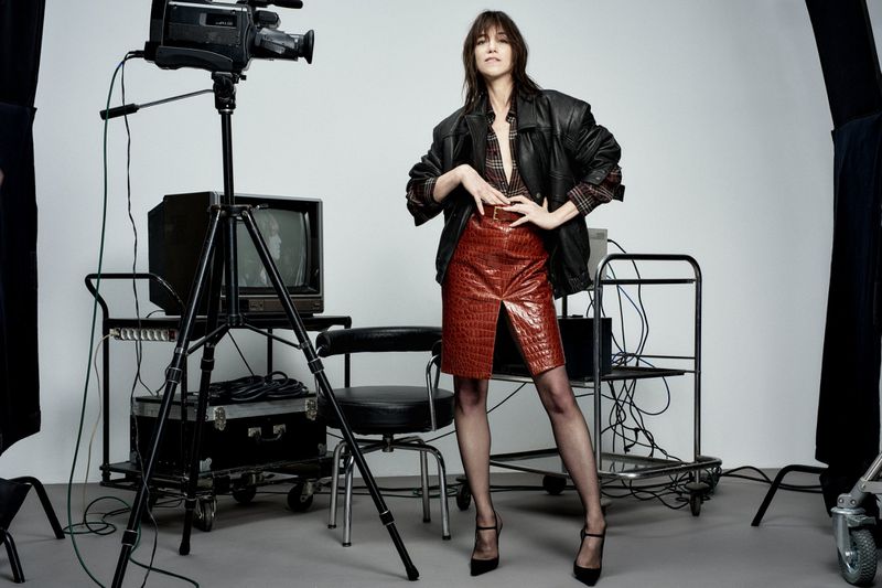 A full-length shot of a woman posing in a studio with vintage video equipment, wearing a leather jacket and red skirt.