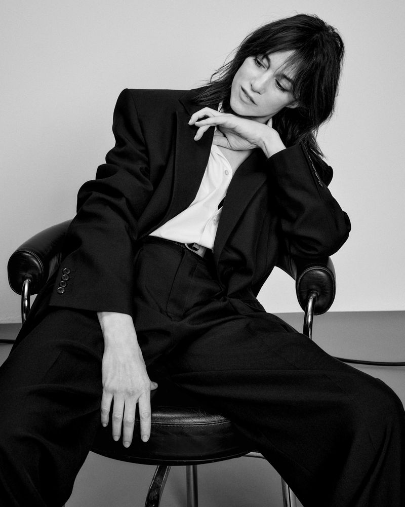 Charlotte Gainsbourg seated in a black blazer and trousers on a dark chair against a plain light background