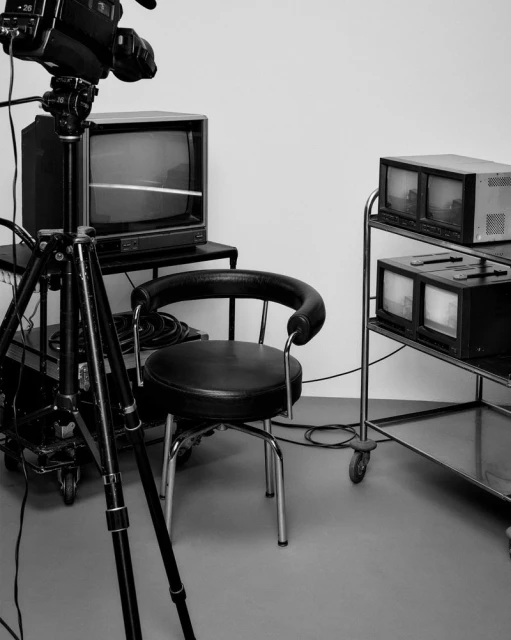 Vintage black and white image of a video camera on a tripod, a vintage TV, a rolling cart, and a black padded chair in a studio setting.