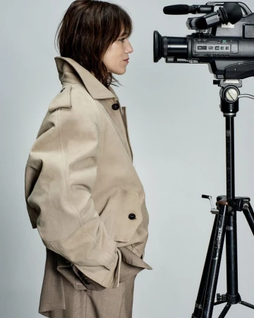 Woman in trench coat looking at large video camera on tripod in neutral indoor setting with soft lighting for visual content creation.