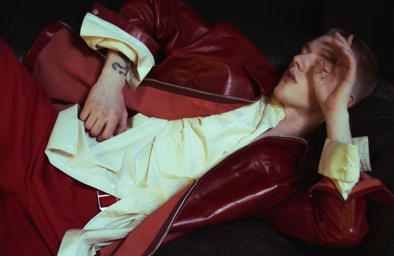 Young man in red leather jacket and matching pants lies on a dark couch with a hand on his face making a ring with his fingers.