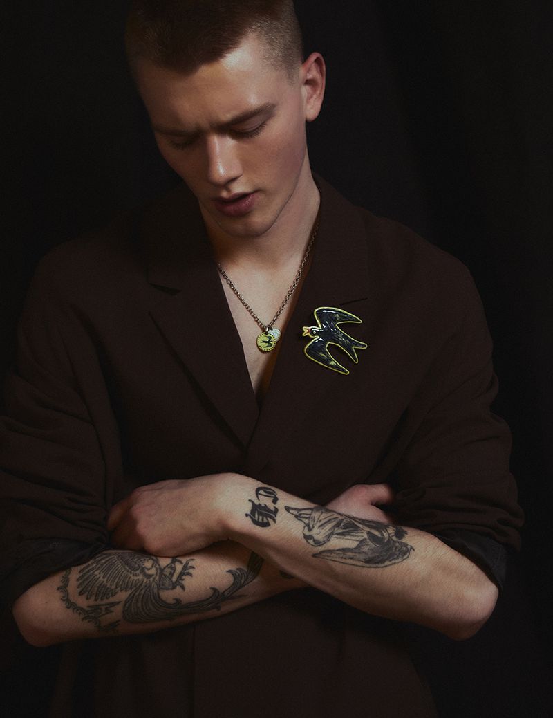 Portrait of a young adult male in a brown jacket with an enameled swallow pin and tattoos on arms against a dark background.