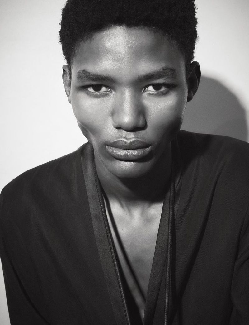 Striking black and white portrait of a young Black male model with soft lighting wearing a casual black shirt against a white background.