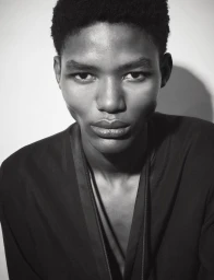 Close-up black and white portrait of a young male model wearing a dark collarless shirt against a plain wall.