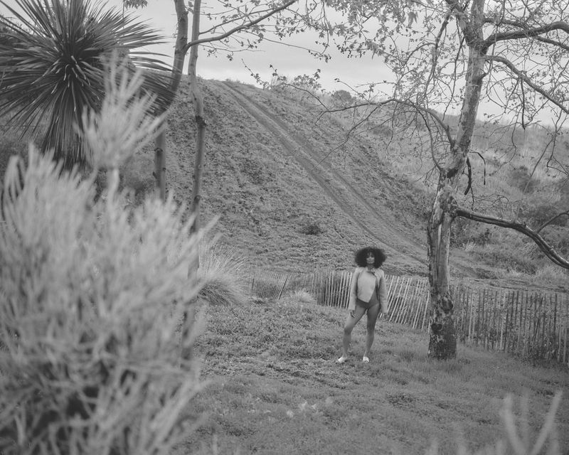 Black and white editorial portrait of singer Ciara standing outdoors on a grassy hillside framed by sparse trees and foliage.