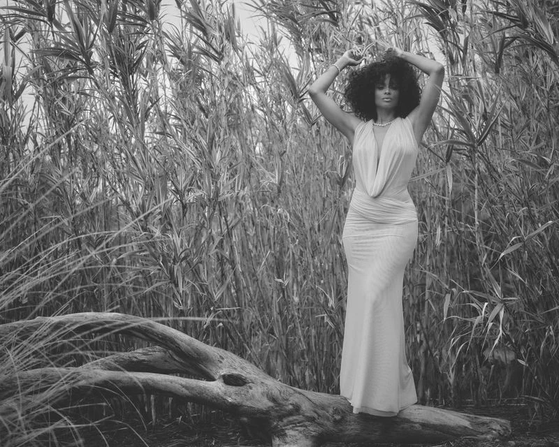 Monochrome portrait of a woman in elegant dress, posing on a fallen tree trunk amidst tall grasses and reeds in a natural outdoor setting.