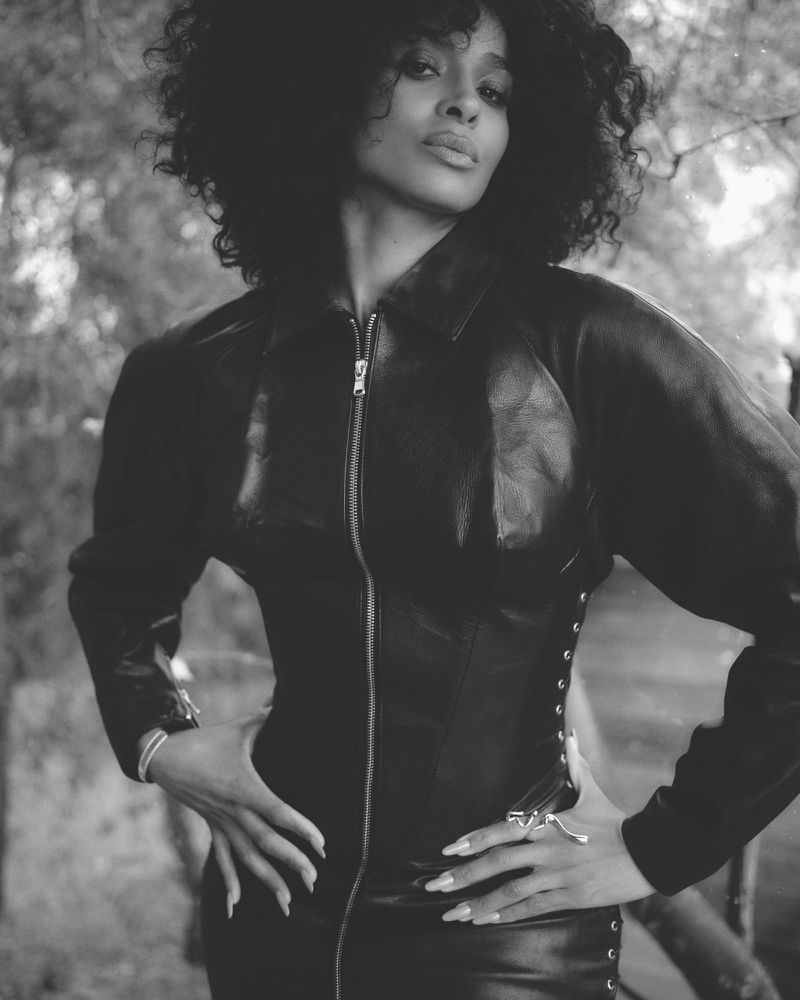 Black and white portrait of Ciara wearing a dark leather zip-up jacket outdoors amidst trees.