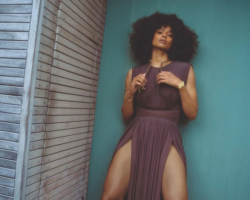 Adult woman with curly hair in a sleeveless purple gown with high slits, posing in front of an turquoise panel and wood slat wall.