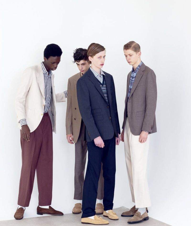 Four young adult men in suits and loafers posing in a studio setting. Three are light-skinned and one is dark-skinned. Clean, bright aesthetic.