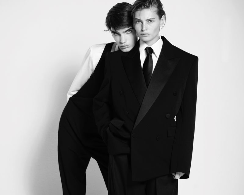 Two stylish models in formal wear pose together against a white backdrop in a black and white fashion-editorial style photograph.
