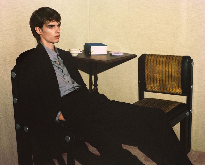 Young man sits in an armchair in front of a wooden table with a book and a cup indoors, modelling in a dark suit and light shirt.