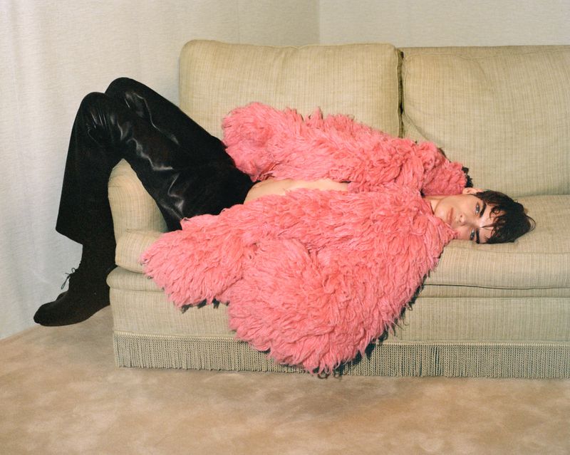 A person lounges on a light-green sofa, clad in black pants, boots, and a fluffy coral jacket, set against a neutral backdrop.
