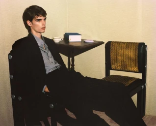 A young man in a dark suit and grey shirt seated on a chair in a corner with a small table behind him.