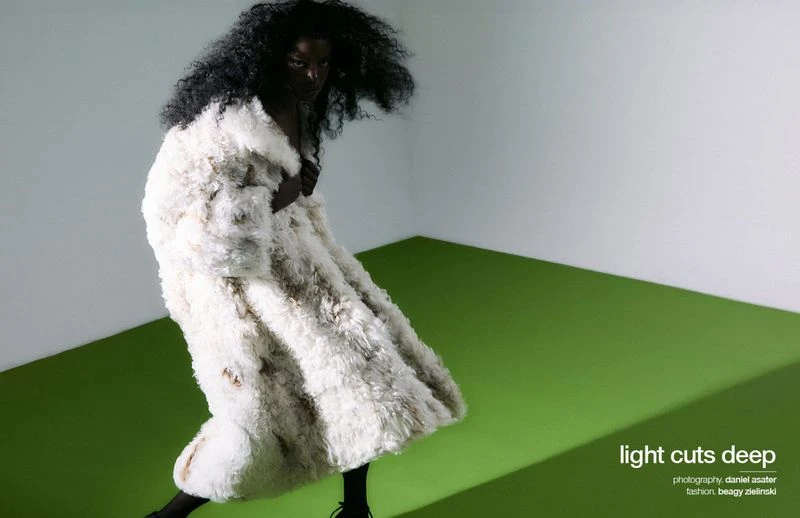 Fashion editorial shot: A Black model wears a textured, off-white coat against a green and white background in a studio setting.